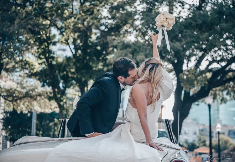Groom kissing the bride as she raises her bouquet from a vintage convertible