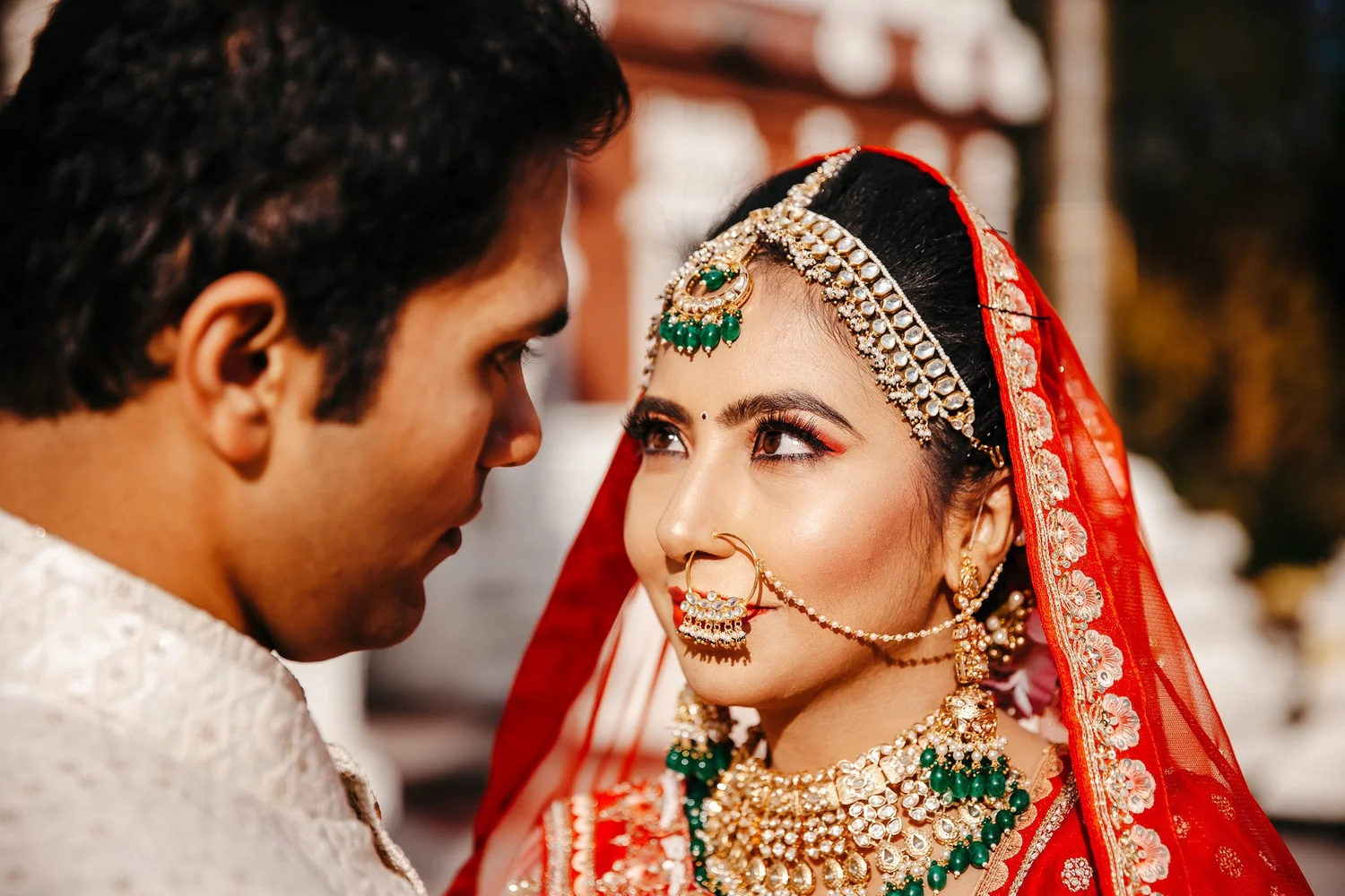 South Asian bride in a red dupatta and gold jewelry gazing at her groom